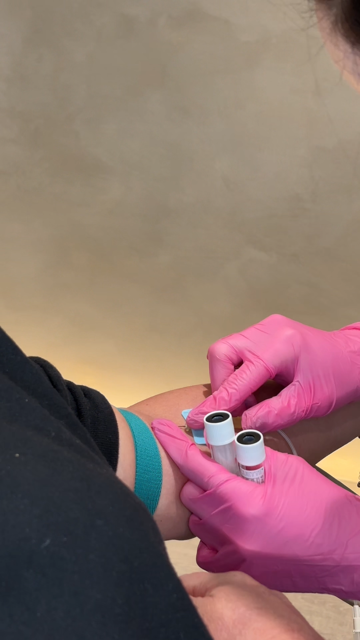 Blood being taken (no needles shown) from arm. Practitioner wears pink gloves; client has a blue strap on arm and wears a grey top.